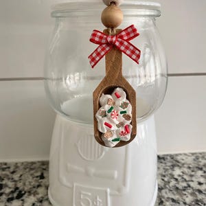 Puede incluir: Una máquina de chicles blanca con una tapa de tarro de cristal transparente. Un adorno de cuchara de madera con un lazo de cuadros rojos y blancos está suspendido desde la parte superior. La cuchara está llena de glaseado blanco y hombres de jengibre y bastones de caramelo en miniatura.