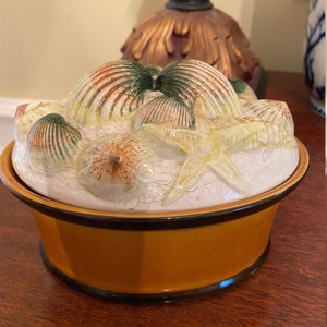 May include: A ceramic container with a lid decorated with seashells and a starfish. The container is glazed in a light brown color with a dark brown rim.