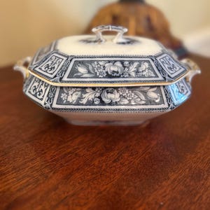 May include: A covered serving dish with a cream-colored base and intricate blue floral and geometric patterns. The dish has a gold trim and handles, and a matching lid with a small handle. The dish is likely vintage or antique.