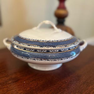 May include: A white and blue ceramic serving bowl with a lid. The bowl features a blue band with gold accents and a decorative pattern. The bowl has two handles and a white pedestal base.
