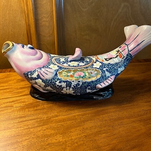 May include: A ceramic fish teapot with a pink and blue glaze. The teapot is decorated with floral and geometric patterns in gold and white. The teapot is sitting on a black wooden base.