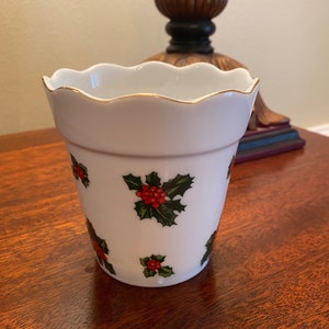 May include: White ceramic vase with a scalloped rim and a gold trim. The vase is decorated with a repeating pattern of red berries and green holly leaves.