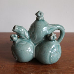 May include: A teal ceramic teapot shaped like three pomegranates. The teapot has a spout and a handle. The teapot is decorated with raised details.