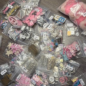 May include: A collection of various rhinestone and metal charms and embellishments in small plastic bags. The charms include pink, silver, and gold colors, with some featuring floral, cross, and logo designs. Some bags have handwritten labels.