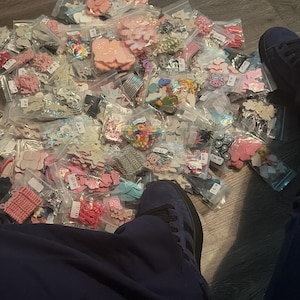 May include: A large collection of small, clear plastic resealable bags filled with various colorful craft embellishments, including shapes like hearts, stars, and flowers in shades of pink, blue, white, and multi-colored patterns, scattered together.