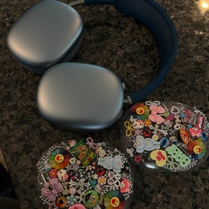 May include: A pair of silver headphones with a blue headband, next to two clear heart-shaped containers filled with colorful charms, beads, and small toys. The charms include a green monster, a pink bow, a yellow smiley face, and a pink heart with the text "BE MINE".