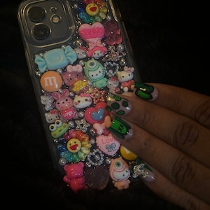 May include: A clear phone case decorated with colorful cartoon characters, candy, flowers, and rhinestones. The case has a camera cutout and a black phone inside.