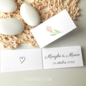 Può includere: Un biglietto pieghevole bianco con una piccola illustrazione ad acquerello di un tulipano rosa sul davanti. L'interno mostra il contorno di un cuore nero disegnato a mano, i nomi Marghe & Marco e la data 04 ottobre 2025 in elegante corsivo.