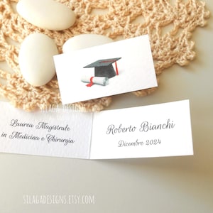 Può includere: Biglietti segnaposto di laurea bianchi con illustrazione di tocco nero e diploma. I biglietti sono piegati, con testo in italiano. Lo sfondo comprende confetti bianchi e un centrino in pizzo beige.