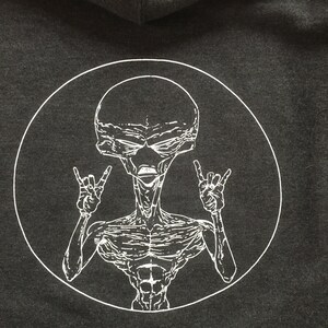 May include: A black hoodie with a white circle graphic of an alien with a large head and a muscular body. The alien is making the rock and roll hand gesture with both hands.