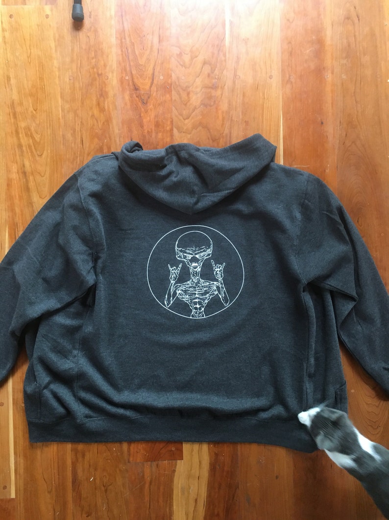 May include: A dark gray hooded sweatshirt with a white graphic of an alien with its arms raised in a rock and roll hand gesture. The alien is inside a white circle.