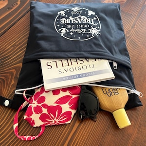 May include: Black zippered tote bag with a white Disney Cruise Line "Treasure" logo. The bag is open, revealing a book, sunglasses, a floral mask, and a bottle of sunscreen. The bag is on a wooden surface.