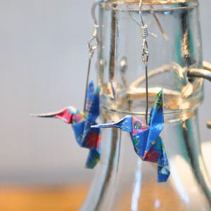 Pu&ograve; includere: Un paio di orecchini origami a forma di colibr&igrave;. Gli orecchini sono realizzati in carta blu con motivi floreali rosa, rossi e gialli. Sono appesi a ganci argentati.