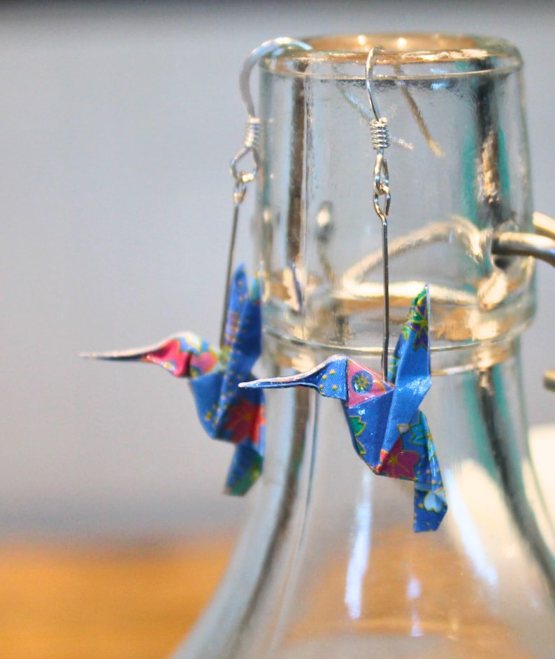 Pu&ograve; includere: Un paio di orecchini origami a forma di colibr&igrave;. Gli orecchini sono realizzati in carta blu con motivi floreali rosa, rossi e gialli. Sono appesi a ganci argentati.