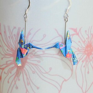 Pu&ograve; includere: Un paio di orecchini a forma di colibr&igrave; origami. Ogni orecchino presenta un colibr&igrave; di carta blu con dettagli floreali, sospeso a un gancio color argento. Lo sfondo &egrave; un motivo floreale rosa e bianco.