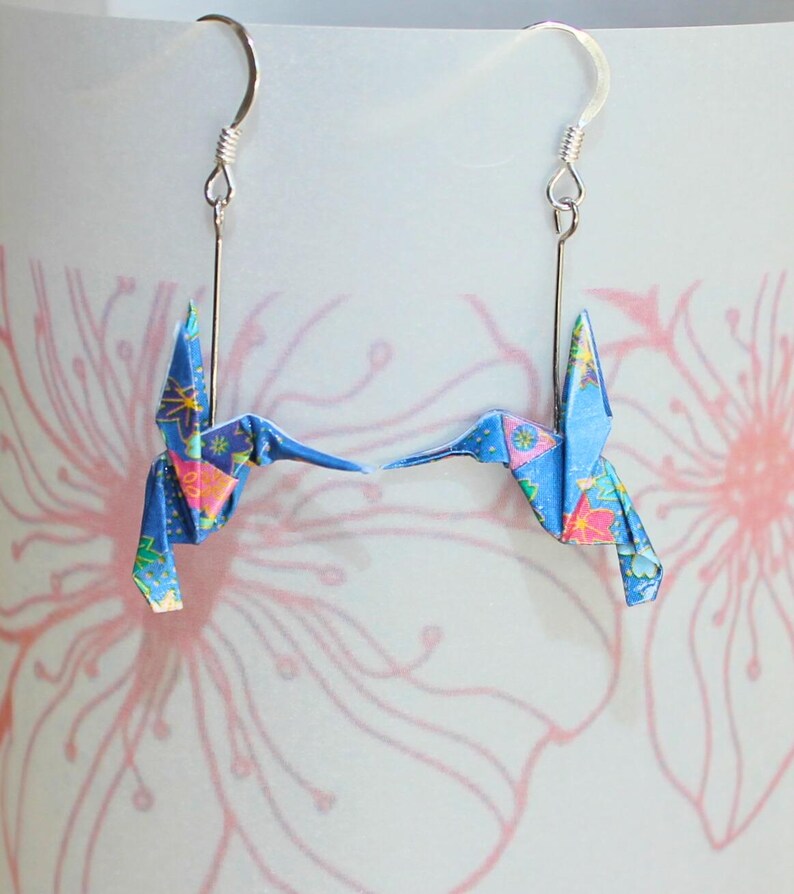 Pu&ograve; includere: Un paio di orecchini a forma di colibr&igrave; origami. Ogni orecchino presenta un colibr&igrave; di carta blu con dettagli floreali, sospeso a un gancio color argento. Lo sfondo &egrave; un motivo floreale rosa e bianco.