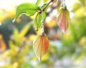 Può includere: Orecchini fatti a mano con un design a forma di foglia. Gli orecchini sono realizzati in carta piegata nei toni del giallo, dell'arancione e del rosa, con ganci in metallo color argento. Sono appesi a un ramo verde e frondoso, su uno sfondo sfocato.