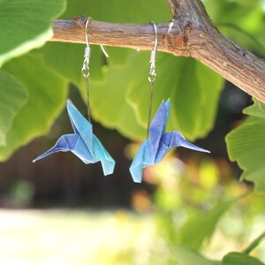Pu&ograve; includere: Un paio di orecchini a forma di colibr&igrave; origami, nei toni del blu e turchese, appesi a ganci d'argento. Gli uccelli di carta sono sospesi a un ramo, su uno sfondo di foglie verdi. Un accessorio unico e fatto a mano.