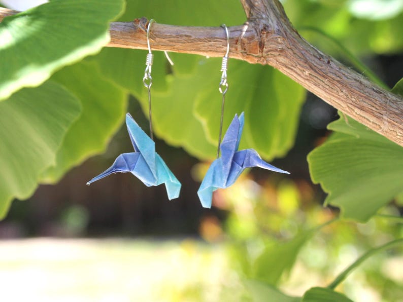 Pu&ograve; includere: Un paio di orecchini a forma di colibr&igrave; origami, nei toni del blu e turchese, appesi a ganci d'argento. Gli uccelli di carta sono sospesi a un ramo, su uno sfondo di foglie verdi. Un accessorio unico e fatto a mano.