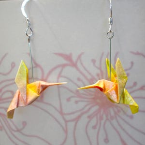 Pu&ograve; includere: Un paio di orecchini a forma di colibr&igrave; origami. Gli uccelli di carta sono piegati in tonalit&agrave; di giallo, arancione e rosa. Gli orecchini pendono da ganci color argento. Lo sfondo presenta un motivo floreale delicato.