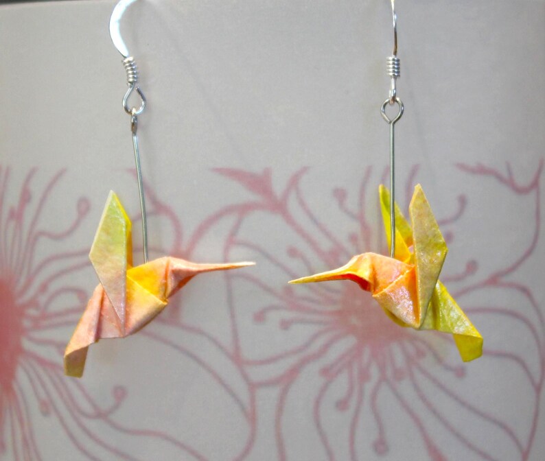 Pu&ograve; includere: Un paio di orecchini a forma di colibr&igrave; origami. Gli uccelli di carta sono piegati in tonalit&agrave; di giallo, arancione e rosa. Gli orecchini pendono da ganci color argento. Lo sfondo presenta un motivo floreale delicato.