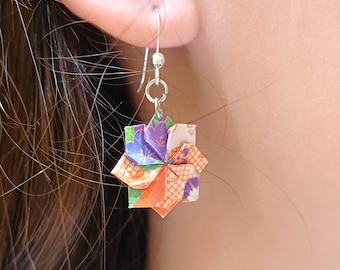 Può includere: Primo piano di un orecchino origami colorato. L'orecchino ha la forma di una stella, con carta piegata nelle tonalità del viola, arancione, verde e bianco. L'orecchino è appeso a un gancio d'argento. Lo sfondo è un orecchio e capelli.