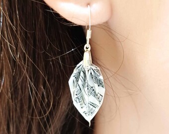 Può includere: Un singolo orecchino con un gancio d'argento e un ciondolo a forma di foglia. Il ciondolo è bianco con note musicali nere stampate sopra. L'orecchino è appeso a un orecchio.