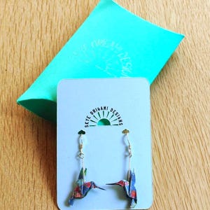 Pu&ograve; includere: Un paio di orecchini origami a forma di colibr&igrave; con ganci color oro, esposti su una carta azzurra. Gli orecchini sono realizzati in carta fantasia con tonalit&agrave; rosse, blu e verdi. Sullo sfondo, una confezione regalo turchese con la scritta "Skye Origami Designs".