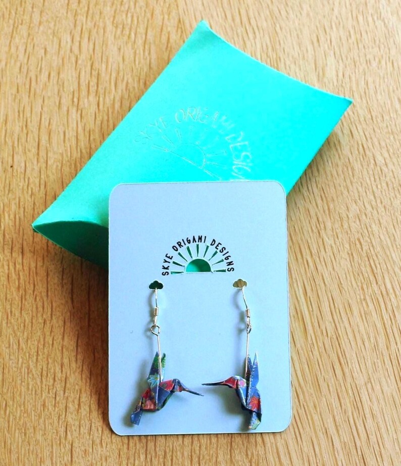 Pu&ograve; includere: Un paio di orecchini origami a forma di colibr&igrave; con ganci color oro, esposti su una carta azzurra. Gli orecchini sono realizzati in carta fantasia con tonalit&agrave; rosse, blu e verdi. Sullo sfondo, una confezione regalo turchese con la scritta "Skye Origami Designs".