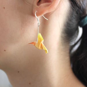 Pu&ograve; includere: Orecchino d'argento con un ciondolo a forma di colibr&igrave; origami giallo e arancione. L'uccello &egrave; sospeso a un gancio d'argento, mostrando l'intricato design di carta piegata. L'orecchino &egrave; indossato all'orecchio di una persona.