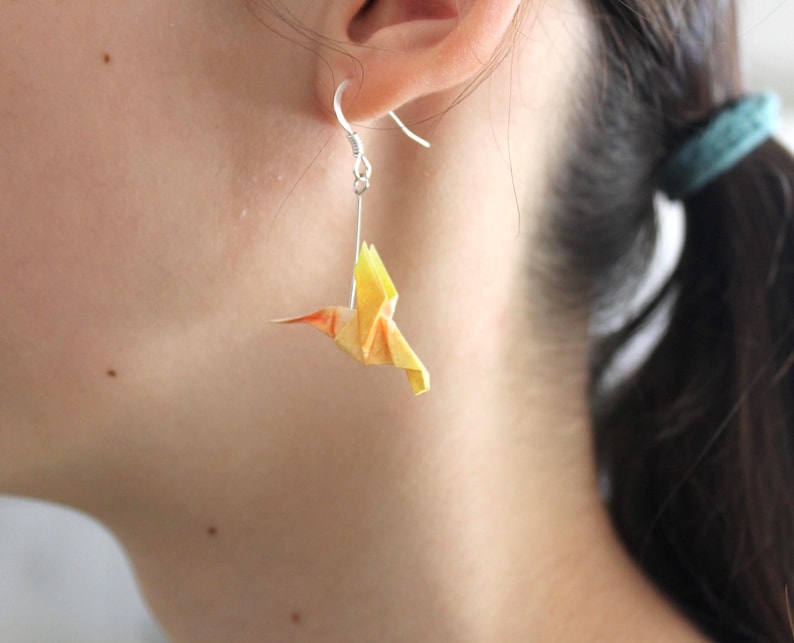 Pu&ograve; includere: Orecchino d'argento con un ciondolo a forma di colibr&igrave; origami giallo e arancione. L'uccello &egrave; sospeso a un gancio d'argento, mostrando l'intricato design di carta piegata. L'orecchino &egrave; indossato all'orecchio di una persona.
