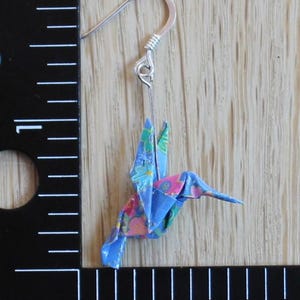 Pu&ograve; includere: Un singolo orecchino a forma di colibr&igrave; origami con un gancio d'argento. L'uccello di carta &egrave; blu con un motivo floreale rosa, verde e bianco. L'orecchino &egrave; esposto su uno sfondo di legno chiaro.
