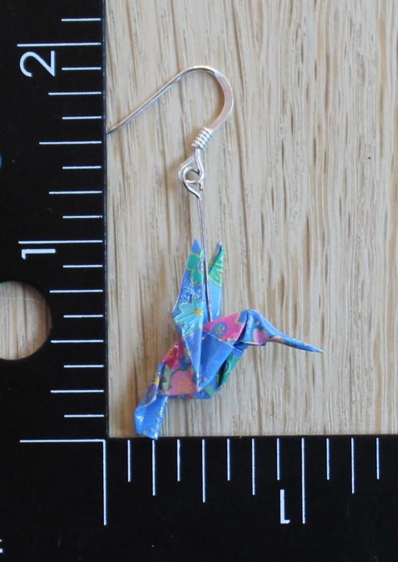 Pu&ograve; includere: Un singolo orecchino a forma di colibr&igrave; origami con un gancio d'argento. L'uccello di carta &egrave; blu con un motivo floreale rosa, verde e bianco. L'orecchino &egrave; esposto su uno sfondo di legno chiaro.