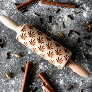 May include: A wooden rolling pin with a carved design of cannabis leaves. The rolling pin is on a grey surface with white flour and spices scattered around it.