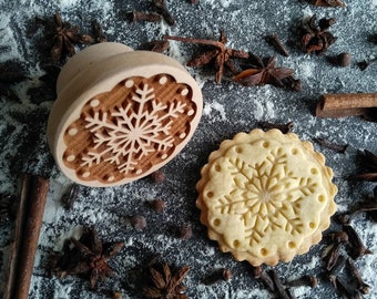 Könnte beinhalten: Ein Holz-Keksstempel mit einem Schneeflockenmuster, neben einem Keks mit dem gleichen Schneeflockenmuster. Der Keksstempel und der Keks befinden sich auf einer dunkelgrauen Oberfläche mit weißem Mehl und braunen Gewürzen.