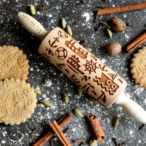 May include: A wooden rolling pin with a nautical themed design. The rolling pin is engraved with images of anchors, ships, and steering wheels. The rolling pin is surrounded by cookies, cinnamon sticks, and spices.