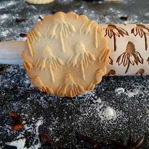 May include: A close-up of a wooden rolling pin with a carved design of flowers. A cookie with the same design is next to the rolling pin. The background is a dark surface with white flour scattered on it.