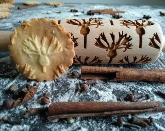 Pode incluir: Um rolo de massa de madeira com um design entalhado de renas com chifres. O rolo está sobre uma superfície com paus de canela, cravos e farinha. Um biscoito com um design de rena está em primeiro plano.
