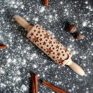 Pode incluir: Um rolo de madeira com um padrão de estrelas e lua. O rolo tem o texto "ROLLING PIN COLLECTION" na pega. O rolo é rodeado por farinha, paus de canela e nozes.