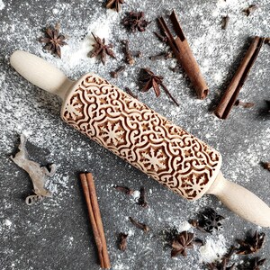 May include: A wooden rolling pin with a carved intricate design, surrounded by cinnamon sticks, star anise, and cloves on a white surface.