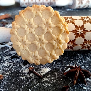May include: A close-up of a snowflake-patterned cookie resting on a wooden rolling pin with a matching design. The cookie is light brown and the rolling pin is natural wood. There is white flour dusting the surface.