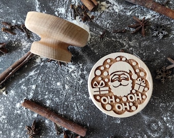 May include: A wooden cookie cutter shaped like Santa Claus with the text "Oh Ho Oh" around him. The cutter is on a gray surface with flour and cinnamon sticks.