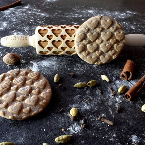 May include: A wooden rolling pin with a heart-shaped design. The rolling pin is on a black surface with a cookie and cinnamon sticks. The rolling pin has the text "Rolling Pin Collection" on it.