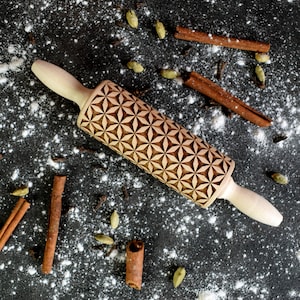 May include: A wooden rolling pin with a geometric pattern carved into the surface. The rolling pin is on a black surface with white powder and brown cinnamon sticks.