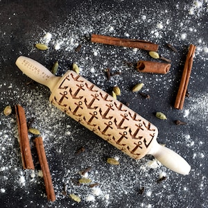 May include: A wooden rolling pin with a repeating pattern of anchors engraved on the surface. The rolling pin is on a black surface with white flour and cinnamon sticks scattered around it. The rolling pin is labelled "Rolling Pin Collection".