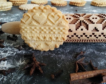 Pode incluir: Um rolo de massa de madeira com um design entalhado de corações e quadrados. O rolo está apoiado em um biscoito que foi enrolado com o rolo. O biscoito é de cor marrom claro e tem um padrão de corações e quadrados. Também há anis estrelado e cravos-da-índia espalhados ao redor do biscoito e do rolo.