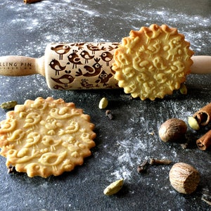 May include: A wooden rolling pin with a design of birds engraved on it. The rolling pin is on a grey surface with a cookie imprinted with the same bird design. There are also cinnamon sticks, cloves, and nutmeg on the surface.