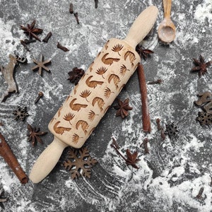 May include: A wooden rolling pin with a carved design of reindeer and leaves. The rolling pin is on a grey surface with flour and spices scattered around it.