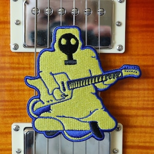 Könnte beinhalten: Ein gelber und blauer gestickter Aufnäher mit einer Cartoon-Figur, die eine Gasmaske trägt und Gitarre spielt, befestigt an einer braunen und goldenen E-Gitarre.