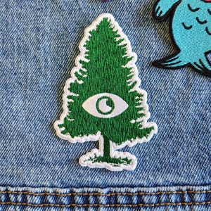 May include: A selection of embroidered patches on a denim background. The patches feature a green tree with an eye, a pink and purple National Park design, and a blue cartoon fish. The denim shows visible stitching.